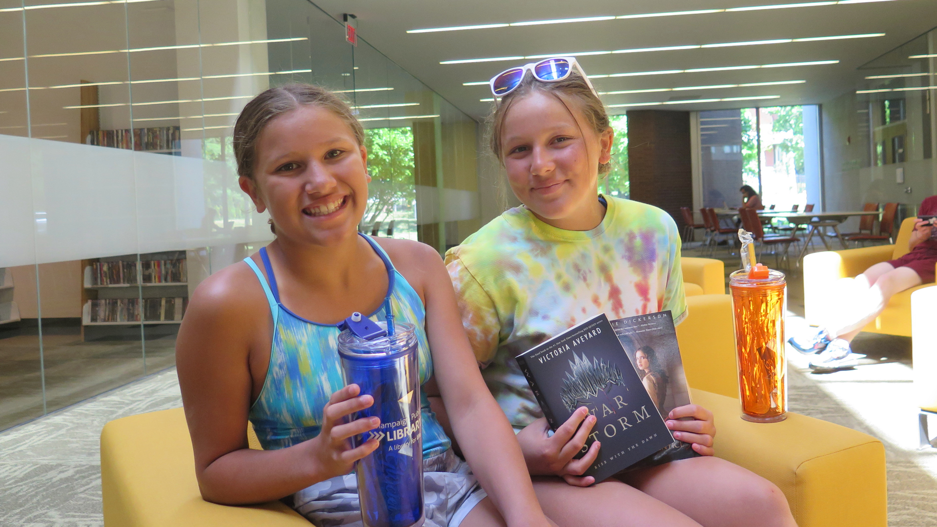 2 teens smiling, holding prize books, tumblers, in library
