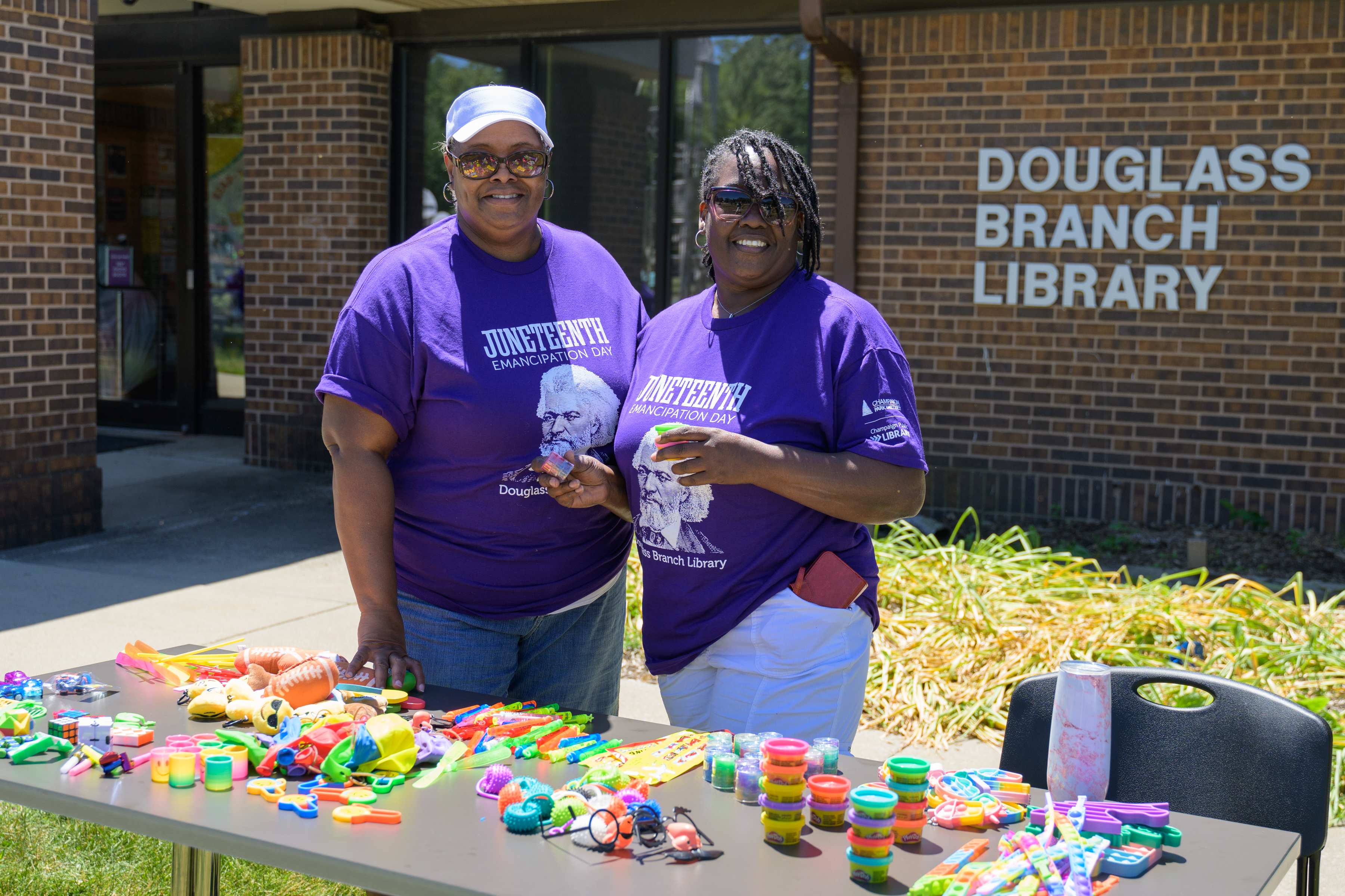 2 volunteers smiling, prize table, in front of Douglass Branch Library