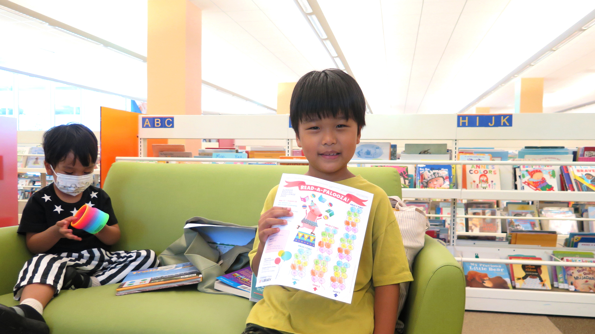 smiling boy holding up colored-in reading log, sitting on couch with toddler in children's area