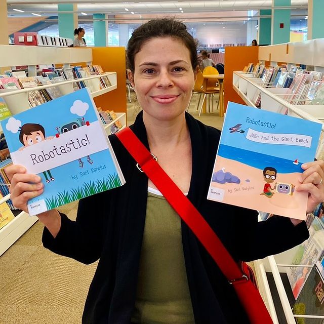 woman holds up 2 picture books, smiling, in children's area of library