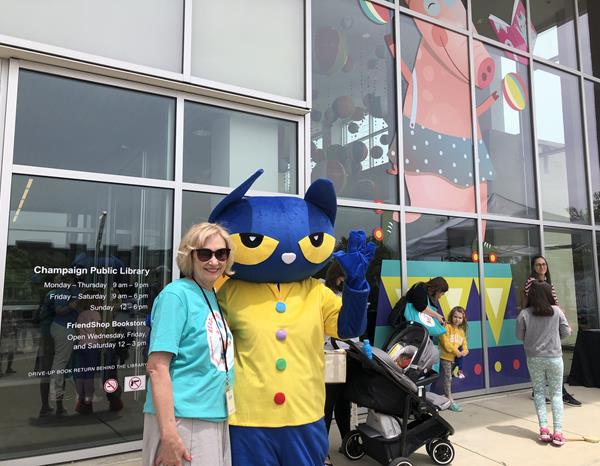 Donna Pittman smiling with life-size Pete the Cat character, front porch of library with families and giant juggling pig graphic behind, on front porch of library