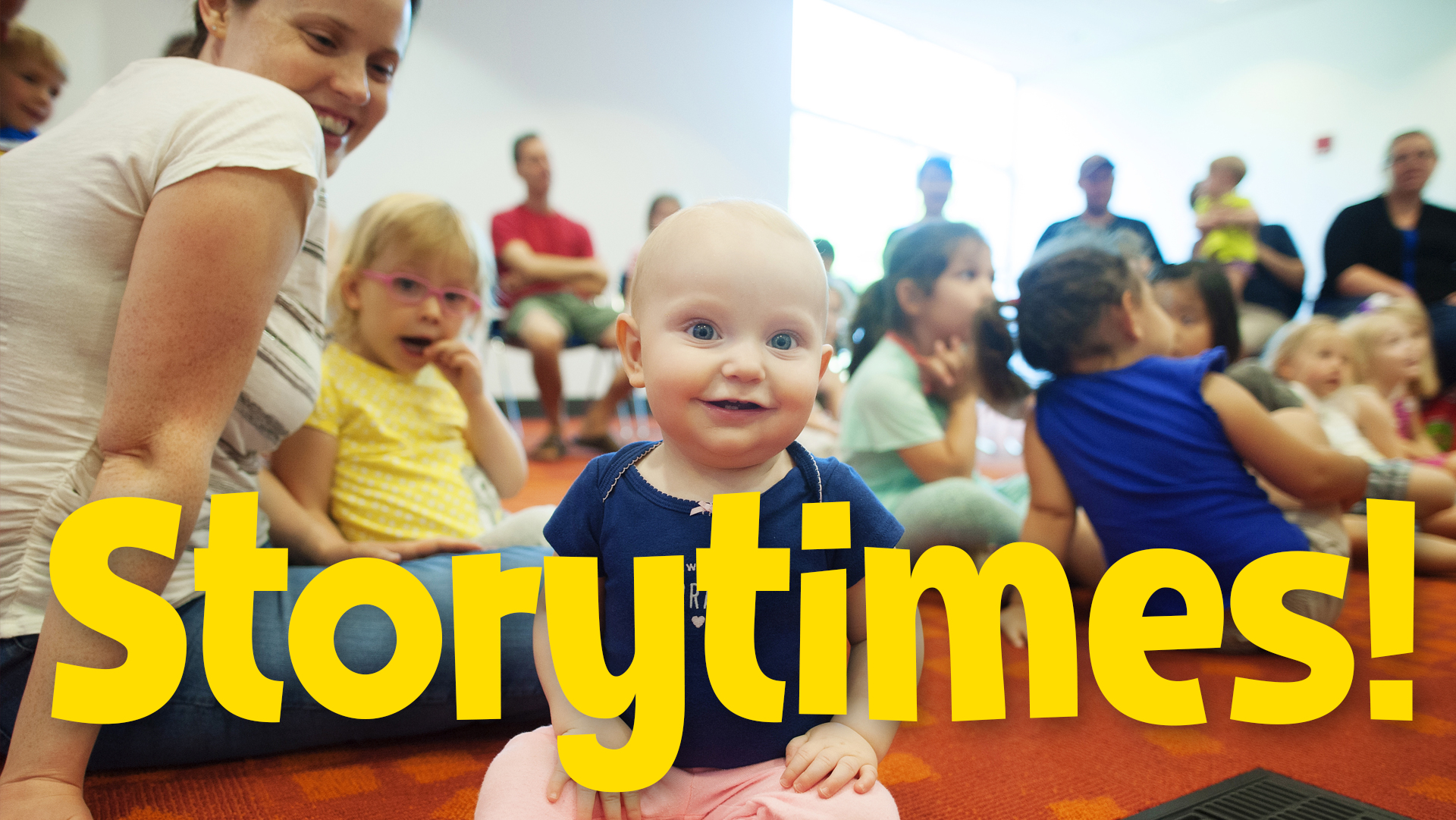 baby smiling at camera, children behind sitting on carpet listening to story, parents in chairs, at the library