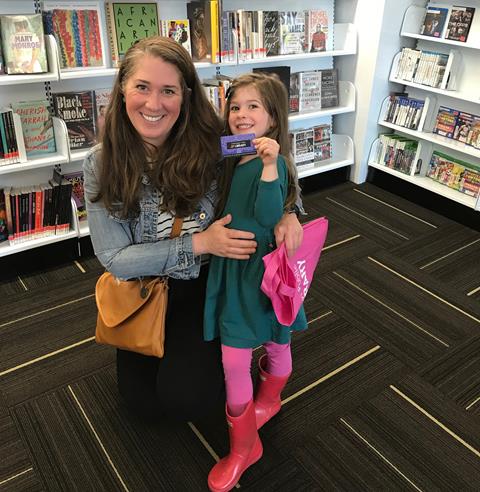 5-year-old holds up new library card, smiling, with mom