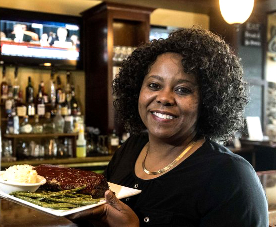 portrait Gayle Starks, holding plate of food, with bar behind at Neil St. Blues restaurant