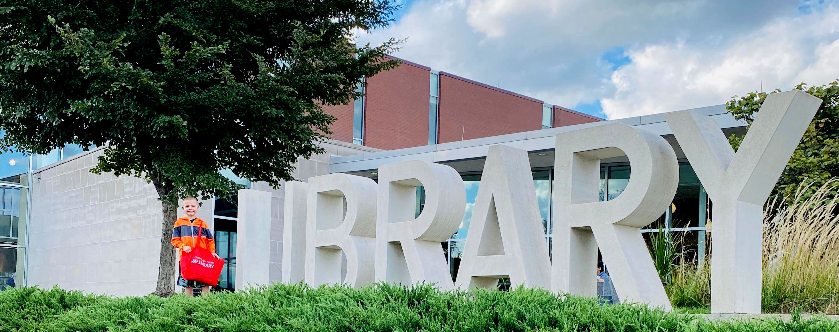 smiling boy stands by LIBRARY sign outside